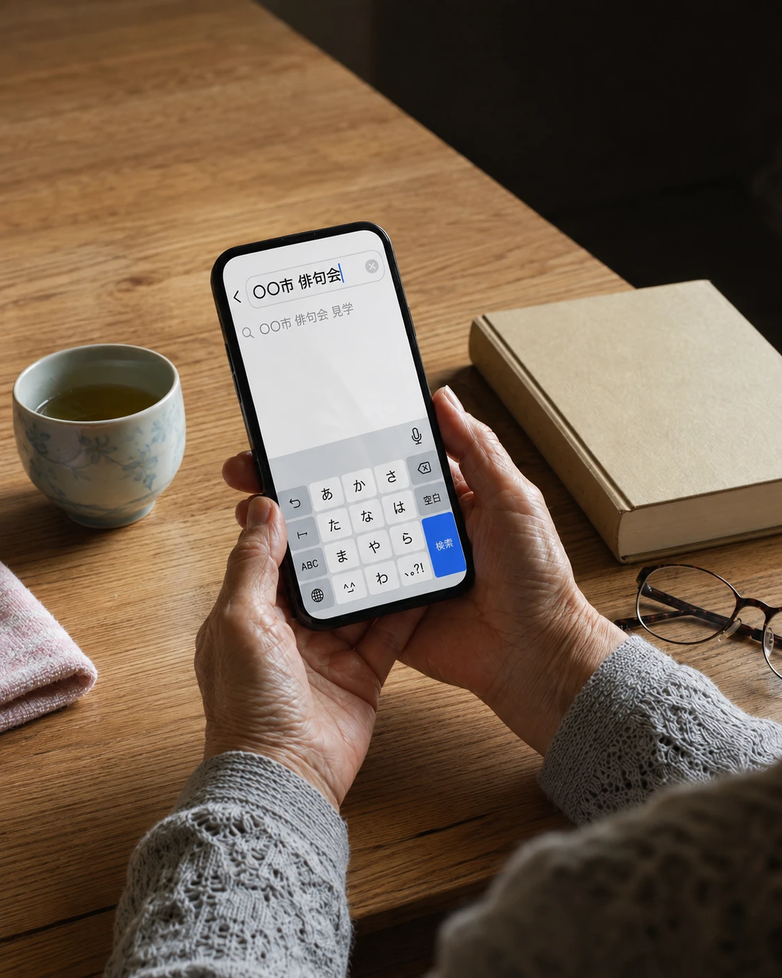 自宅の食卓で、老眼鏡と本を傍らに、スマートフォンで「〇〇市 俳句会」と検索している年配の人